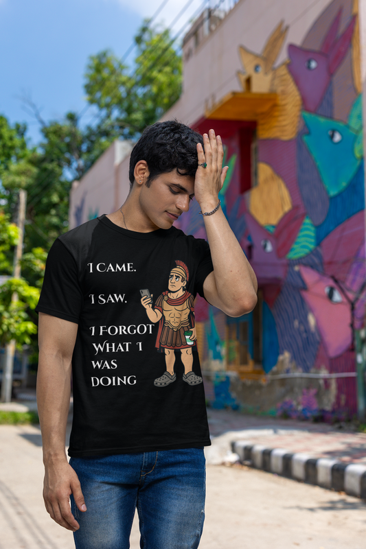 a man wearing a black I Came, I Saw, I Forgot T-Shirt – Funny ADHD t shirt from WhimSpin with text and graphics, standing in front of a colorful mural. he has his hand raised to his forehead as if he is just remembering something he forgot.