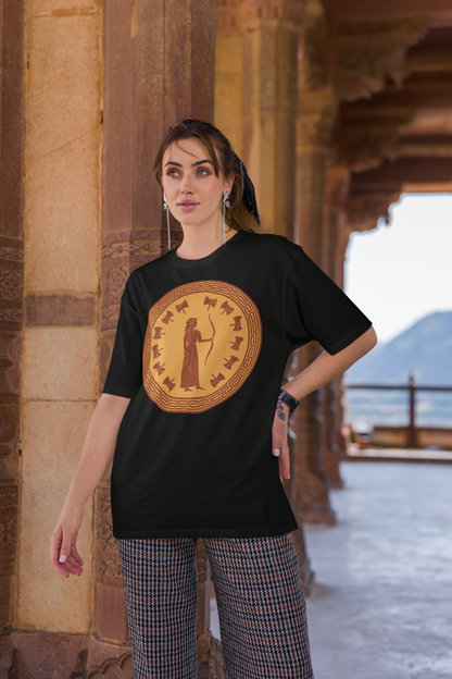 A woman with dark hair stands with her left hand on her hip. She stands in an ancient building and is wearing the Penelope Epic Queen of Ithaca T shirt in black from Whimspin. The shirt features an image of a woman in ancient Grecian robes holding a bow and surrounded by 12 axes. The images is in terracotta cotta red and gold in the style of ancient amphora art. 