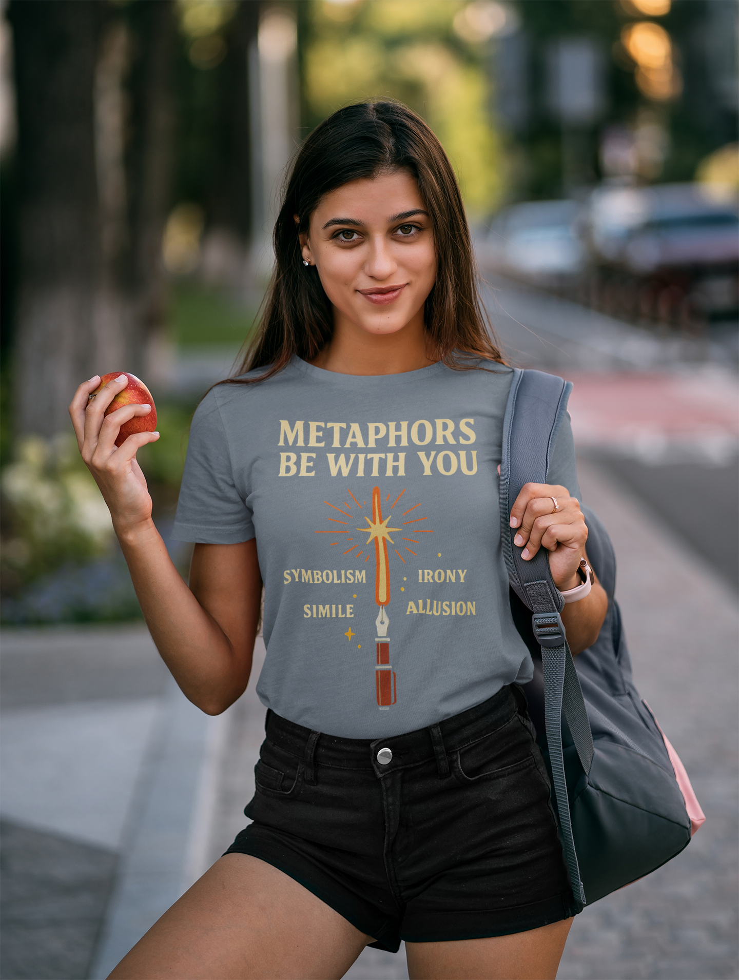 a college aged woman wearing a black Metaphors Be With You T-Shirt – Inspired by Sci-Fi & Lit Class Heroes shirt from WhimSpin standing outdoors on a city street holding an apple in her right hand and carrying a backpack over her left shoulder. The graphic features a fountain pen with a ray of light coming from the nib like a light saber and the words "Metaphors Be With You" over top 
