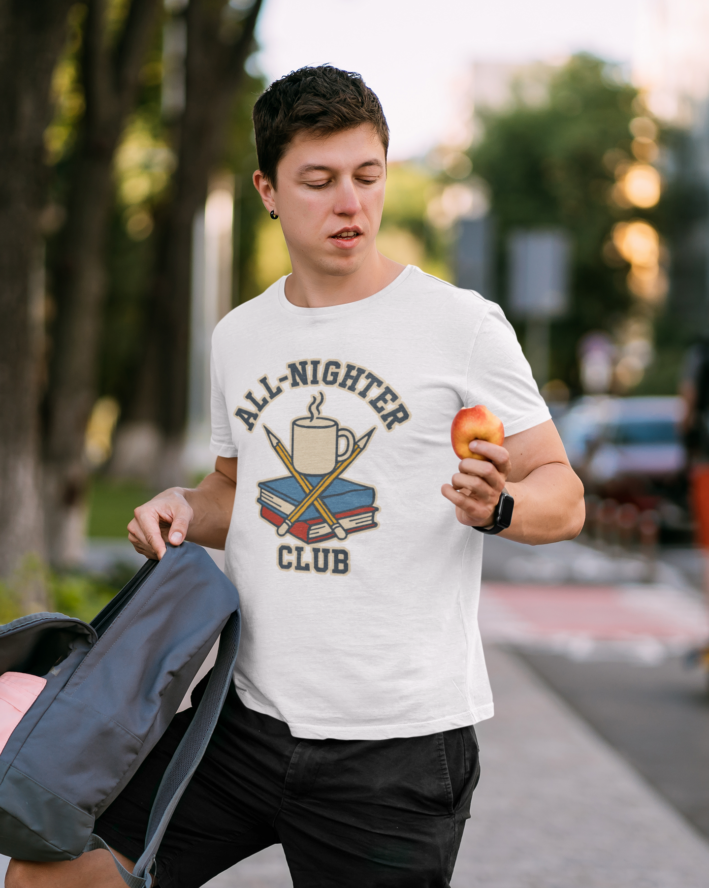 Person wearing a white All-Nighter Club T-Shirt – Funny Caffeine & Study Tee for Night Owls shirt from WhimSpin with 'All-Nighter Club' design, holding an apple outdoors.