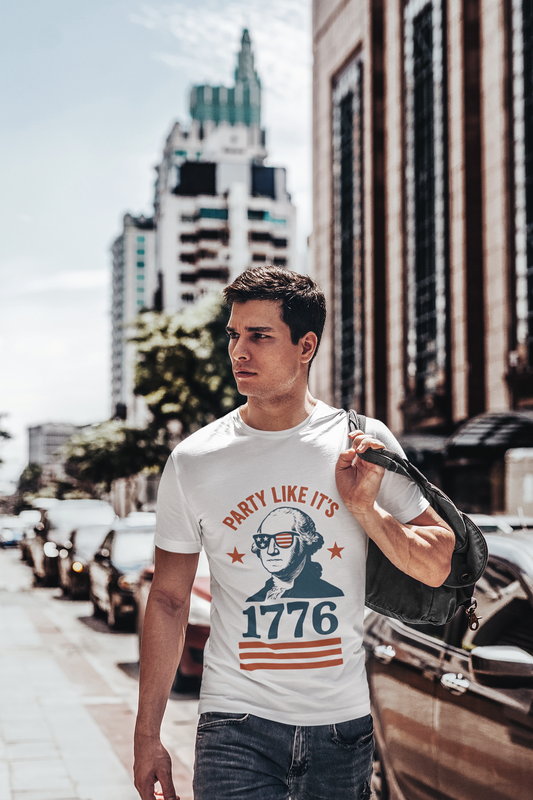 A caucasian man with brown hair wearing a funny white Party Like It's 1776 shirt with George Washington wearing slitted US Flag sun glasses walks down a city street with his coat thrown over one shoulder. 