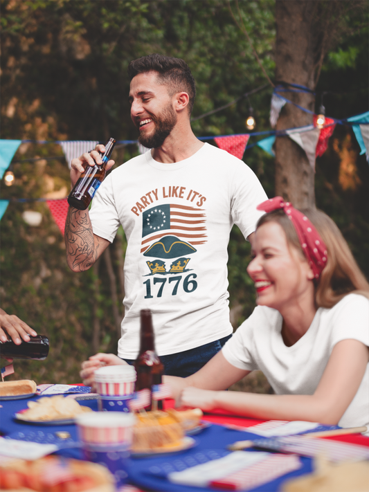A man with dark hair wearing the Party Like It’s 1776 T-Shirt – Revolutionary Spirit USA Tee from WhimSpin stands next to a table with red white and blue 4th of July party decorations while raising a bottle of beer towards his mouth. The shirt graphi features the words "Party Like It's 1776" a 13 star colonial flag and a tricorner hat sitting on-top of a crown.